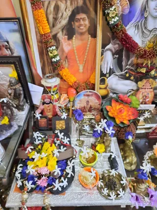 Photograph of a home shrine set up for daily worship. A central framed portrait shows a smiling man in orange robes with a long beaded necklace, raising one hand in greeting, with marigold and rose garlands draped around the frame. Beside it on the right, a large image of a seated Shiva figure with a white body and hand raised is also garlanded. On a silver tray in front are a pair of black padukas (sandals) decorated with yellow and blue flowers and jasmine, small bowls of offerings, a brass deity figure, a kalasha, a conch shell, and an orange lotus arrangement. A small seated figure in red and gold stands on the left. No people are physically present.