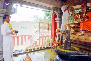 A photograph of a temple shrine during an abhishekam ritual. On the left, a man in white dhoti with a yellow shawl and rudraksha mala holds a brass oil lamp and stands before a row of brass kalashas set along a wet stone floor. Beside him, a second man in white dhoti and yellow shawl stands on a raised step with hands folded in prayer, facing a golden deity murti seated on a lotus base and draped in orange cloth. In front is a black stone shivalingam on a round yoga-peetha basin, with red flower petals scattered nearby. A red curtain hangs behind, and a tiled roof and green trees show through an open pillared doorway.