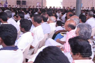 A photograph of a large seated audience at an outdoor event, viewed from behind and at the side. Rows of white plastic chairs are filled with men mostly in white shirts and white dhotis or trousers, with some in pastel or checked shirts. Many have short dark hair, and several bald or greying heads are visible. A speaker stands at upper left and a stone-textured wall with a banner forms the backdrop. The crowd fills the frame and extends into the distance.