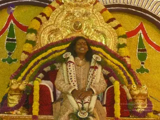 A smiling person with long dark hair sits on an ornate gilded throne decorated with thick garlands of yellow, orange, white and purple flowers arranged in arches around the seat. They wear a pale cream and gold embroidered sherwani-style outfit with white and red-tipped flower garlands draped over the shoulders and chest, and their hands rest folded in their lap. Gold lion-head armrests flank the red-cushioned seat, and the backdrop is a flower-covered yellow wall with red and green decorative motifs and a gold sunburst frame above. The scene resembles a ceremonial seating honouring a guru, with the central figure appearing as an unsure candidate for a person based on the throne setting and ceremonial clothing alone.