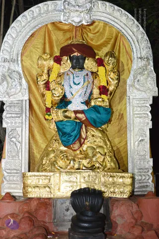 A black stone deity murti is enshrined in a silver-framed arched niche against a gold draped backdrop. The figure wears a maroon turban, a teal and gold garment, layered white and silver jewelled necklaces, and a gold armband, with yellow and red flower garlands draped over the arms. Multiple arms hold gold attributes, and a dark stone lotus-shaped base sits in the foreground on a red stone platform. No physical people are visible; the scene is a night-time shrine display.