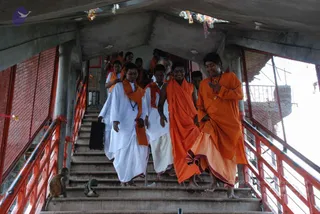 A group of men in saffron and white robes descends a concrete stairway between red metal railings and wire mesh, with small festive strings of ribbons hanging from the ceiling above. The front man on the right, in a long orange robe with a rudraksha mala, has a swept-back hairstyle and gestures with one hand; the man beside him wears an orange shawl and gestures similarly. Several others in white dhotis with orange shawls follow, including one carrying a black bag. Two monkeys sit on the steps at the left edge.