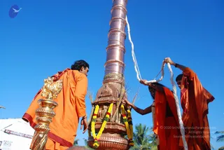 Outdoor flag-hoisting ceremony under a clear blue sky beside a tall copper-clad flagstaff. Two figures in orange ochre robes stand at its base: a man on the left in orange with a maroon-edged shawl, bent forward and hands low near a gold lion-headed post; a figure in the centre in orange with long dark hair, one hand on a garlanded stone shrine-like base decorated with marigold and yellow garlands and sheaves of grass, the other arm raised to the white rope. A third figure on the right in orange robes pulls the white hoisting rope above. A small stone deity murti sits within the base. Palm trees and a banner with Kannada text appear at the left edge.