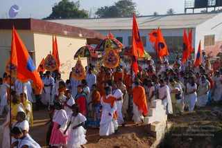 An outdoor procession moves along a dirt path past low buildings under a clear sky. Many devotees walk in a crowd, many in white clothing. Tall orange flags with a blue circular emblem are carried upright, and several decorated umbrellas with multicoloured canopies and gold-fringed tassels are held aloft. In the centre a man in an orange shawl and a woman in a red sari walk with others; a man in white dhoti and orange shawl walks near the front, and girls in white dresses with pink or white skirts and beaded necklaces walk in the foreground. A small white stone structure sits at the right edge beside the path.