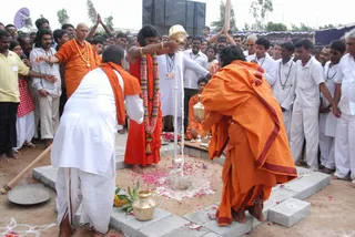 Outdoor ritual at a construction site with a square brick-lined pit. A bare-headed man in an orange robe with a red-bordered cloth pours liquid from a brass pot through a white pipe into the pit, while a man in orange with a long shawl bends over it with a small vessel. A third man in orange-red robes wears a thick red and white flower garland and gold bangles and bends at the pit's side. Standing nearby are a shaved-headed man in orange with a beaded necklace and a man in white with a red-bordered cloth and white garment, both with palms pressed together. Around the pit are scattered rose petals, a brass pot and a brass lamp. Behind, a crowd of men and boys in white shirts and white dhotis with mala beads stand watching under a tent, with a dark banner and a blue-and-white flag visible.