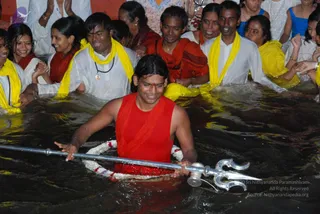 A photograph of a night water ritual in a pool. In the centre, a man with long dark hair and a red vertical tilak on his forehead stands waist-deep in dark water, wearing a sleeveless red garment and a white flower garland at his waist. He holds a polished silver trident upright in his right hand and smiles. Behind him, devotees wearing white clothes and bright yellow shawls stand in the water with their hands raised in prayer. Several women in red and white saris and a man in an orange cloth also stand among them, some clapping. A bright orange-red wall with a small purple logo is visible at the top edge.