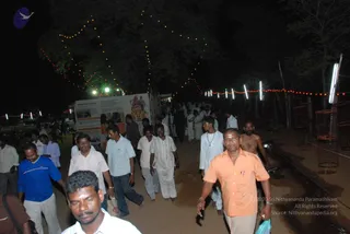 A night-time photograph of a crowd of men walking along a dirt path lit by strings of fairy lights and red bulbs strung through trees. Most are in white or light shirts and dhotis or trousers; one man in front wears an orange shirt and another in a blue top walks to the left. A shirtless man stands on the right beside a row of tall white light poles. On the left a white signboard with orange borders and illustrations is set up beside the path. No identifiable a person candidate is visible in the frame.