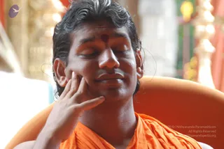 Close-up photo of a man seated in an orange armchair, eyes closed, one hand resting against his cheek. He wears an orange saffron wrap, has a red bindi-style tilak mark on his forehead and short, slightly tousled dark hair. Behind him are blurred gold and white decorative pillars and red fabric. The pose is calm and contemplative.