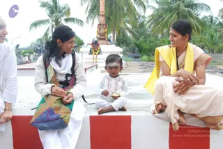 A photograph taken outdoors at a temple grounds with palm trees behind. Three women and a toddler sit on a white ledge edged with red-and-white painted bands. On the left, a woman in a white dress and white scarf with a shoulder bag of multicoloured patterned fabric smiles toward the centre. In the middle, a small toddler in a white outfit with red embroidery and a white-and-red forehead mark holds a small brass dish. On the right, a woman in a cream sari with a yellow shawl, a red bindi and a vermilion-marked parting smiles while resting her hands on her knee. Behind them stands a white plinth with a garlanded stone sculpture of a seated bull, a tall copper-coloured pillar and a garland of marigolds, with carved stone figures nearby. A partially visible woman in white stands at the far left edge.