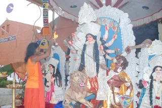 A photograph of a standing man in an orange saffron wrap and long dark hair lifting a flame in front of a large white-lace deity murti of Durga, a ten-armed goddess with a crown and a lion mount. A golden-clad figure with a sword stands beside the goddess, and smaller white-lace deity figures flank the scene. A brass oil lamp rests in the foreground, and a sign reading Ananda Ishwar Mandir hangs on a brick wall behind. The setting suggests an aarti before a deity under a decorated canopy. The man has long, swept-back hair and orange clothing, a cue consistent with a person's presentation, though his face is partly turned away, so the identification is uncertain.