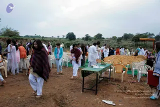 An outdoor gathering on bare red earth in a rural field with scrubby trees and a cloudy sky. Rows of yellow and white plastic chairs stand on green mats, and a small green-draped table holds a tray with offerings. Many people in white and pastel clothes walk or stand about; a woman in the foreground wears a dark patterned shawl over white trousers, and a girl in a pink top and maroon shawl walks barefoot. A woman in a turquoise jacket and lanyard is near the chairs. At the right edge an orange-canopied structure stands beside a table with flowers and decorations.