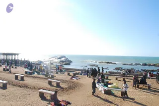 A sandy seaside promenade under bright sky beside a calm sea with rocky outcrops. Groups of people stand and sit on the sand near white concrete benches; some spread colourful cloths and bags on the sand. A small open pavilion with pillars stands at left. A man in white walks with a bag at right and a few people in dark clothing stand near the water's edge. A pedestrian cycles along the edge. No identifiable a person is visible; the scene shows devotees and visitors gathered by the shore.