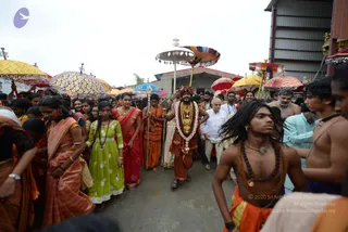A photograph of an outdoor procession on a paved path with a corrugated metal building behind. In the centre, a man wearing a rust-orange garment, a long orange-red headwrap, a heavy gold necklace and a thick white and pink flower garland walks forward holding a staff, with a shaved-headed attendant beside him in white. Around them, women wear colourful saris and salwar in red, orange, green and yellow, many with gold jewellery, and devotees in orange and saffron clothing carry a long staff and walk among decorated umbrellas in gold, purple, red and blue. On the right, a young man with long dreadlocks and a bare chest wears rudraksha beads and an orange cloth. The centrally placed man is an appearance-based a person candidate, marked by his flower garland, gold necklaces, saffron-orange clothing and head covering, leading the procession.