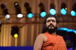 A man with long dark hair and a full dark beard stands on a stage, looking upward. He wears a sleeveless saffron-orange top and a matching orange shawl draped over one shoulder, and has a white tripundra marking with a red kumkum dot on his forehead. A small headset microphone is attached near his face. Behind him, blurred stage lights in blue, white and yellow glow against warm wooden panelling. The figure has distinctive long swept-back hair, saffron clothing and a shawl, and the page indicates Swamiji attended this event; appearance-based identification is probable.