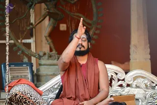 A photograph of a bearded man seated on an ornate silver carved sofa with eyes closed, right hand raised with palm facing forward near his forehead in a blessing or initiation gesture. He wears a rust-orange shawl over a maroon top and orange lower garment, with a red thread on his wrist and white sacred markings on his forehead. Behind him a bronze statue of a dancing deity stands against a deep red wall, with a stone pillar on the right. A leopard-print cushion and an orange cloth lie beside him, and a silver-framed sofa edge is visible. Two other people appear only partially at the bottom edge. Swamiji (probable): long dark hair, rudraksha-style attire and orange ceremonial clothing, seated as the presenter of a satsang-style initiation session.