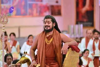 A man with long dark dreadlocks and a full beard sits with eyes closed and a slight smile, wearing a rust-orange shawl draped over his shoulders and a rust-orange robe, with a gold multi-strand necklace and a gold pendant. He rests his right arm on a golden throne-like seat with a red cloth folded over its armrest. Beside him stands a gold trident-topped staff and a golden bird-headed ornament. Behind him, seated devotees wear white garments with orange-red shawls, and a painted image hangs on the wall. A purple and white bird logo appears in the upper left. This appears to be a discourse or satsang setting; the seated man is Swamiji (probable), identified by his long swept-back hair, saffron-orange robe and shawl, and ornate gold necklaces.