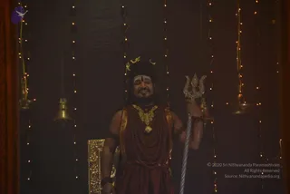 A man stands in a dimly lit room hung with strings of small warm lights. He wears a crescent-moon crown with a tilak on his forehead, a heavy rudraksha mala, a large gold ceremonial necklace, and a maroon-red wrap and dhoti, with rudraksha bracelets on his wrists. He holds a silver trident in his right hand and smiles. Brass bells hang on the left, and a gold-framed panel rests beside him. The figure's distinctive adornments and trident, with no clear face-based identification, present an appearance-based candidate of a person, though the cues are mixed, so identity is uncertain.