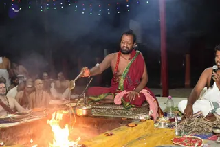 A homa fire ritual at night beneath strings of multicolored fairy lights. A bearded man in a deep red dhoti-style garment with a green and gold border and a pink shawl sits on an ornate silver seat, pouring liquid from a long ladle into a fire pit with bright flames. He wears a red rudraksha mala and a red forehead mark. Around him, several bare-chested priests sit in white dhotis with rudraksha malas, and a man in white clothes and a white shirt holds a microphone and sits beside a brass plate of flowers and dried sticks. A copper vessel, a silver lamp, and a red-and-white floral offering tray lie on the yellow platform.