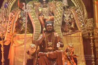 A man with long swept-back dark hair, a white tilak on his forehead, and a rust-orange robe sits cross-legged on an ornate golden throne with lion-head armrests. He wears multiple rudraksha malas and a large ceremonial gold pendant, and raises one hand in a blessing gesture. Behind him rise tall golden backrests studded with white gems, flanked by two large gilded deity murti, one with a trident-like staff standing in front. A golden carved pillar and brass lamps frame the stage. Swamiji (probable) is seated centre on the throne, with the wearer's long hair, rudraksha malas and ceremonial gold pendant as visible cues.