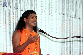 A photo of a man with long dark hair and a small red bindi, wearing a sleeveless saffron-orange garment and a long gold-beaded necklace with green and gold beads. He smiles broadly, one hand pressed to his chest, speaking into a silver microphone on a flexible stand. Behind him hangs a backdrop of vertical strands with orange and red beads. a person appears as the speaker, with long swept hair, saffron clothing and ornate beaded necklace, seated mid-discourse.