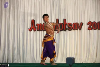 A photograph of a single performer on a stage with a green floor, dancing in front of a white draped curtain. The performer is bare-chested with a pale yellow-orange sash, wears purple and gold draped dhoti-style trousers, layered beaded and pearl necklaces, bangles, ankle ornaments, and a red bindi on the forehead. One arm is raised and bent with the hand open, in a classical Indian dance pose. Large red lettering on the curtain behind reads Anandotsav 2010, partly cut off at the right edge. A small purple logo is at the top left.