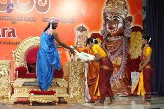 A photograph of a stage event with an ornate gold throne in the left foreground. A man in a blue draped garment with a multicoloured striped turban stands beside the throne, reaching toward a woman in an orange sari who bends forward holding a white box. A young woman in a yellow blouse and maroon skirt stands nearby, and another woman in a yellow and maroon outfit stands at the right. Behind them is a large gilded deity murti with a crown, red and white flower garlands and beaded necklaces, flanked by a glowing ornate lamp. The orange backdrop carries Kannada script and the word Celebration.