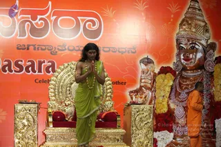 A standing figure with long dark hair, wearing a green sari-style draped garment with a gold border and a rudraksha-style bead necklace, presses palms together in a namaste greeting in front of an ornate gold throne with red cushions. Behind, a large orange banner with Kannada script and a golden deity murti adorned with garlands, jewelry and a crown fills the right side. A smaller figure in orange robes stands at the right edge beside the murti. Flower garlands and a small brass offering tray sit near the throne.