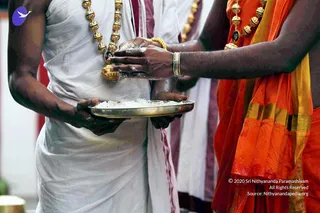 Close-up photograph of a ritual in progress. On the left, a man in white dhoti and white upper cloth wears a long necklace of golden beads with a dark pendant, and holds a brass plate heaped with white powder. On the right, a person in an orange-saffron garment with gold-bordered cloth and gold bangles extends hands dusted with white powder over the plate. Both carry the same white powder on their fingers. The scene suggests a puja or sacred offering, and the attire suggests a temple setting.