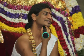A man seated in front of a backdrop of floral garlands in bands of white, purple, red and yellow, with gold ornamental metalwork behind. He holds a black microphone with a teal grip near his chin and looks off to the side with a slight smile. He wears a white unstitched cloth draped over one shoulder, a heavy gold multi-bead necklace across his chest, and a small red mark on his forehead. His hair is long and swept back into a knot. The figure appears to be a person seated for a discourse, with appearance cues including long swept-back hair, ceremonial gold necklace and a seat in a garlanded setting.