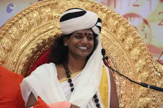 A smiling person sits on an ornate gold throne with carved floral scrollwork and a red cushion, wearing a white turban with black trim, a red bindi, a white shawl over a white garment with gold border, and a long rudraksha mala. A microphone on a stand is near the face. Orange cloth is visible at the lower left and a second orange garment at the bottom. Behind the throne is a pink-and-gold backdrop with partial lettering.