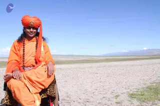 A photograph of a man seated outdoors on open, stony high-altitude ground with a pale lake and snow-capped mountains in the distance under a clear blue sky. He wears an orange turban with a round ornamental brooch, an orange robe and shawl, and a strand of dark rudraksha beads, with a leopard-print cloth draped over his lap. His hands rest on his knees and he smiles toward the camera. Swamiji (probable): orange turban and robes, rudraksha mala, and long hair mark him as a sannyasi-style figure seated among Kailash-Yatra scenery.