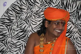 A smiling man sits against a black-and-white zebra-striped backdrop, looking off to the side. He wears an orange saffron turban with gold edging, an orange shawl draped over his shoulder and chest with gold border, and a red bindi on his forehead. He has long dark hair falling behind his shoulders and wears a thick ceremonial gold necklace of beads.