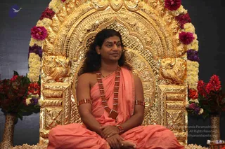 A seated man in saffron-orange robes sits cross-legged on an ornate gold throne with carved lion-like side motifs and a gold arched backrest. He wears a long rudraksha mala with a ceremonial gold pendant, matching rudraksha armlets, and a red bindi on his forehead, with long swept-back hair. His hands rest folded in his lap. The throne is framed by garlands of white, yellow and magenta flowers and tall floral arrangements on either side. Swamiji (probable): the long hair, saffron robes, rudraksha mala and ornate gold throne are consistent with the page's description of the event.