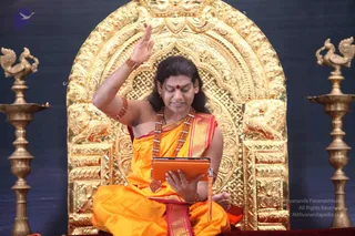 A seated figure in orange and gold silk clothing sits before an ornate gilded throne with carved peacock-like motifs. He wears a rudraksha mala with a long strand and a red bindi, raises one hand in a gesture while looking down at an orange tablet held in his other hand. Brass oil lamps stand on either side of the throne against a dark blue backdrop. Swamiji (probable): the appearance cues are long swept-back hair, an ornate gold necklace with rudraksha beads, saffron clothing and a golden throne, consistent with a discourse setting.