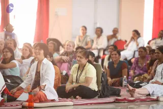 A large hall with pale peach walls and red curtains holds a seated audience on red-and-white striped floor mats and chairs. In the foreground, a smiling woman in a white shirt and orange skirt sits cross-legged beside a younger woman in a yellow top and black trousers wearing a red bindi and beaded lanyard. Behind them, many attendees in white, pink, purple and blue saris and kurtas sit attentively, some wearing mala necklaces. The scene appears to be a satsang or discourse session.
