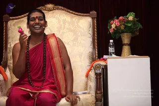A smiling man in a red-pink robe with a gold-bordered shawl sits in an ornate gilded chair with cream and gold floral upholstery against a dark red drape. He wears a long rudraksha mala and a red bindi on his forehead, and holds a small pink object in his raised hand. A wireless microphone is clipped near his face. Beside him stands a white lectern, a clear water bottle, and a gold woven vase of pink and cream flowers.