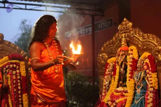 A person with long dark hair in an orange-saffron dhoti and shawl, wearing a rudraksha mala and gold bangles, holds a lit oil lamp aarti flame near two deity murtis adorned with yellow and red marigold garlands and gold jewellery. A stone-pillared temple wall behind them bears a sign reading Sri Nanjundeshwara Mandir. a person: the figure shows long swept hair, saffron clothing and rudraksha mala, consistent with the page's identification of the event, but the face is not used for identification.