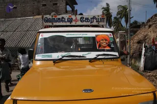 A yellow vehicle viewed from the front in a village street with thatched huts and palm trees behind it. A blue and white roof sign reads Venkatesh Keriya in Tamil script with a small emblem. The windscreen carries a sticker with a green Tamil heading and a framed photo of a smiling man with a shaved head wearing an orange-saffron turban and orange clothing with a vermilion forehead mark, labelled as a sticker on the vehicle. Two windscreen wipers are visible, and a few people stand at the roadside to the left.