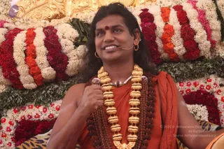 A photograph of a smiling man seated in front of a backdrop of red, orange and white marigold garlands and green foliage. He wears an orange-saffron robe draped over one shoulder, a long multi-strand gold necklace with a rudraksha mala beneath it, and a red bindi on his forehead. A small clip-on headset microphone is attached near his ear, and he holds a microphone near his chest while speaking. Gold-and-leopard-patterned fabric is visible at the lower edge. The appearance matches the Swamiji (probable) cues of long swept-back hair, saffron clothing and rudraksha malas, shown in person as a speaker at a satsang-style gathering.