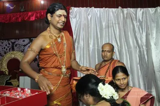 A photograph of an indoor room with dark wood panelling, a red valance and a white satin curtain behind. A standing man in saffron-orange dhoti and shawl, wearing layered gold necklaces, gold armlets and a gold waist belt, with a tilak on his forehead and a rudraksha mala, stands at left with his hand extended toward a seated woman in a red-and-white sari who is wearing a jasmine flower in her hair. Nearby, a bald man in orange clothes and a rudraksha mala stands behind a tray. At left a red tray holds small packets and a gold-and-red ornate chair is visible.