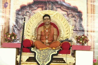 A man seated cross-legged on an ornate gold throne with a red cushioned back and a tiger-skin rug, smiling and wearing orange ochre robes, a wrap over one shoulder, multiple rudraksha malas and ceremonial gold pendants, gold armbands and a tilak mark on his forehead. A long staff leans against the left side of the throne, and pink and white flower arrangements sit on red-topped side tables. Behind him is a gilded decorative backdrop.