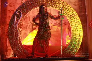 A figure with long dark curly hair stands on a red-draped platform inside a large ornate golden ring-shaped frame with embossed panels and a carved serpent-like arch on the left. The person wears a patterned, leopard-print style garment over a red dhoti-like wrap, with multiple beaded necklaces and mala strands across the chest, bangles on the arms, and holds a tall green-tinged trident (trishul) in one hand, smiling with a dramatic stance. Warm red and yellow stage lighting and a yellow cloth backdrop surround the figure, with a background of carved temple-style relief panels. The image is a photograph of a staged performance-style scene.