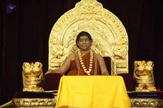 A seated figure sits on an ornate gold throne with a large arched back carved with floral and scroll patterns and two gold lion-head armrests. He wears orange-red robes draped over one shoulder, a heavy gold necklace and a rudraksha-style bead mala, with a red bindi on his forehead, and a small microphone clipped near his face. A yellow cloth covers a table in front of the throne, and dark red velvet drapes form the backdrop. a person: the figure wears orange robes, a rudraksha-style mala and ceremonial gold necklaces, seated on a gold throne, consistent with a satsang setting.
