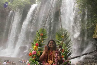 A smiling person with long dark hair, wearing a rust-orange garment, a red bindi and layered flower garlands with a gold ceremonial pendant, sits on a flower- and palm-leaf-decorated swing suspended by ropes in front of a large waterfall. The person raises one hand in blessing and holds a curved brass horn. Several people bathe in the pool below the falls, and dense green foliage frames the scene. The appearance matches the Swamiji cues of long swept-back hair, saffron-toned clothing, rudraksha-style garlands and ceremonial gold necklace, so this is a probable Swamiji (probable) seated on the swing as the central figure.