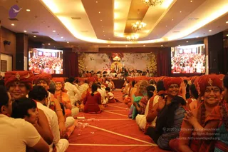 Wide view of a large hall with a red carpet and rows of seated devotees on the floor. Foreground women wear red and orange turbans with a gold emblem and smile, some with orange and red saris. Men in white kurtas sit among them. Two large screens show the crowd, and a central stage has a floral backdrop and a decorated throne-like seat with a figure in white at its centre, flanked by small lamps. Gold chandeliers hang from the ceiling.