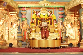 A photograph of a seated man on an ornate golden throne (peetha) in a richly decorated temple-style hall. He wears a maroon-red turban, a red-brown dhoti-style garment and a red garland with a gold pendant, and he holds an open printed booklet or photo-card with a picture. Behind him is a large floral backdrop of yellow, orange and red marigold garlands framing a golden canopy with a kalasha finial. Golden lion-shaped armrests and gilded pillars with sculpted figures flank the throne; standing gilded deity figures stand on either side, one holding a trident. Oil lamps stand on brass lamp stands at the sides. Red carpet covers the floor, with stage lights in the foreground. Several people are visible in the scene.