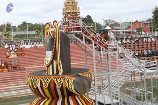 Outdoor ceremony at a large decorated Shivalinga draped in colourful flower garlands with orange, white, purple and green bands, set in a water pool beside red-brick steps. A metal staircase climbs to a raised platform where several saffron-robed priests stand near a gold-topped shrine tower with a kalasha. On the left, a row of white-clad devotees sits on the steps, and at the bottom left, seated figures in saffron robes sit near a small seat with a parasol. Spectators in white and light clothing sit in tiered rows on the right, and a red-roofed building and palm trees stand in the background.
