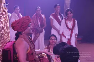 A seated man in a red-maroon turban and a red-orange garment with a heavy gold-coloured beaded necklace sits on a golden throne-like seat at left, smiling and extending his hand toward a devotee who bows with hands folded in front of him. Behind them, several women in white garments with red-and-white shawls stand in a purple-lit hall, one holding her hands together. The seated figure shows distinctive ceremonial adornments and a turban consistent with the page's reference to a person, so this is a probable identification in the appearance-based sense; the devotee and standing women are shown in darshan-style setting.