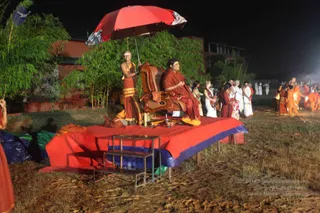 Night outdoor photo of a ceremony on a raised platform covered with a red cloth with blue edging. A seated person in a rust-red robe sits in a large brown leather armchair, with a red and white umbrella held over them by a shaved-headed attendant in a rust-red and gold-bordered dhoti with a gold necklace. Brass ritual objects and a small metal step stool sit on the platform. Behind them, bamboo stands by a red-brick building, and a crowd of people in white, red and orange robes stands and moves around the grounds with flickering lights in the background.