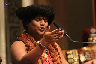 A man with long dark hair piled high on his head and a red tilak on his forehead speaks into a boom microphone, one hand raised in a gesture. He wears a thick necklace of large rudraksha beads and a gold ceremonial pendant, a coral-pink garment, and a beaded bracelet. Behind him, a blurred figure in a red and gold ornate costume sits in the background. The setting is an indoor hall with warm lighting.